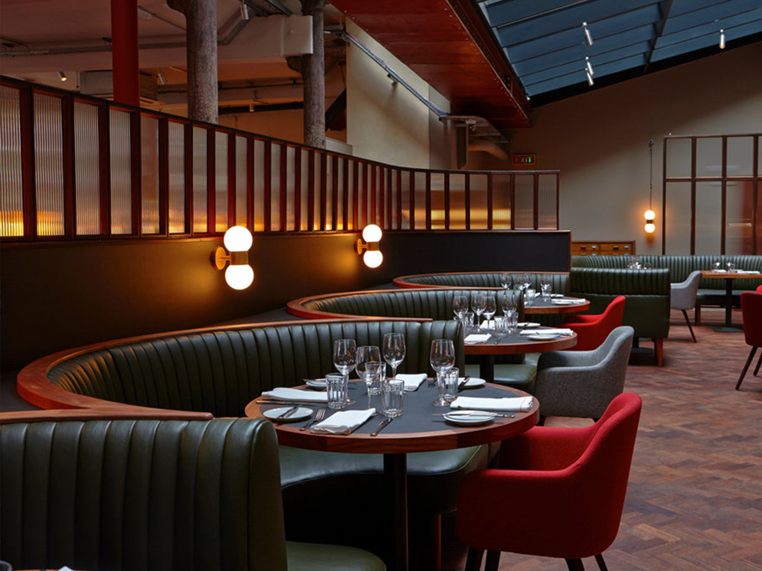 A modern restaurant interior with round tables, set for dining. Green leather booths, red and gray chairs, wooden floor, and wall lamps create a stylish, contemporary atmosphere.