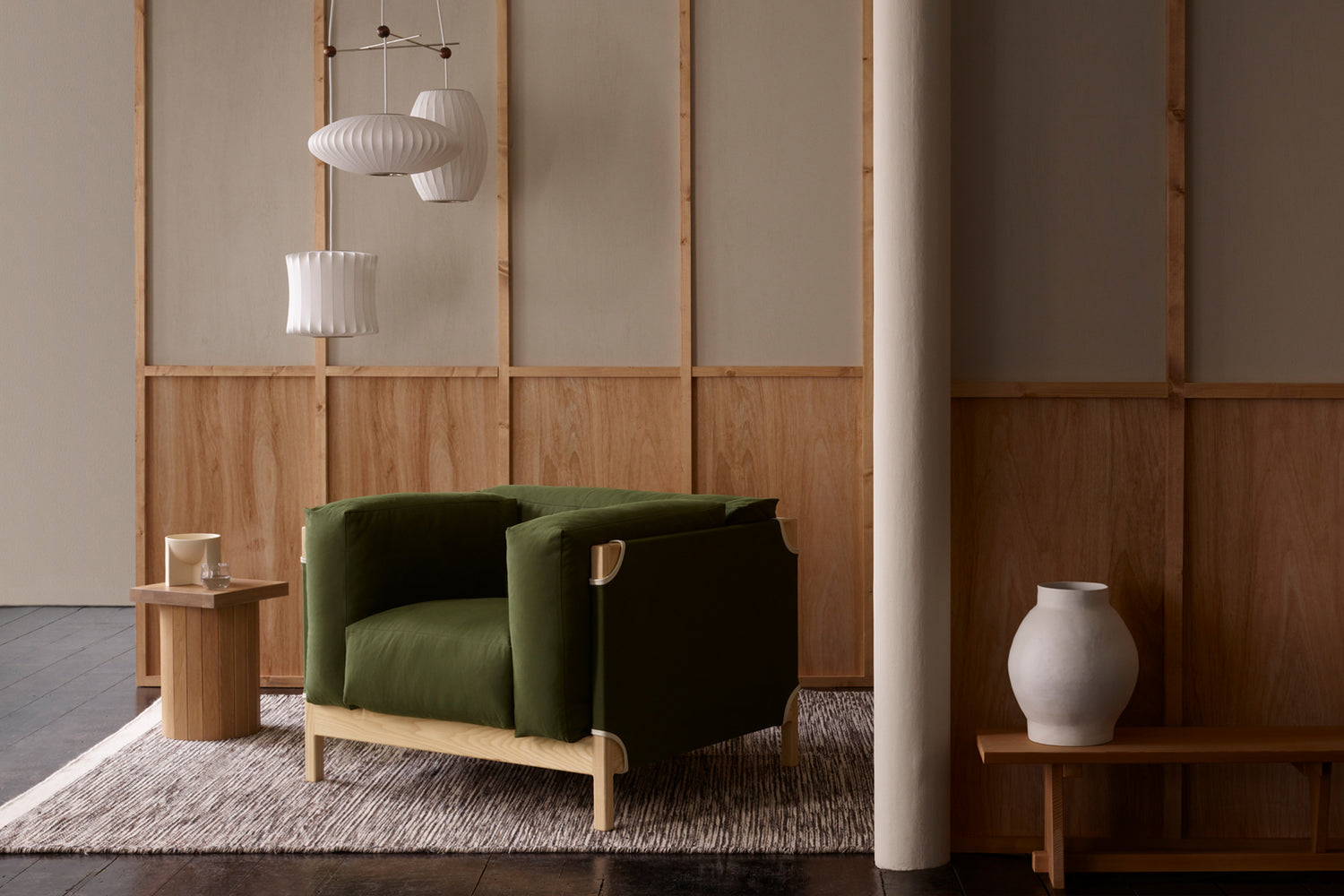 A green armchair with wooden frame sits on a rug beside a small table and white vase, with pendant lights hanging in a minimalist, wood-paneled room.